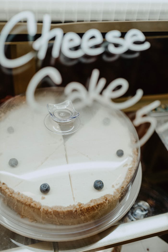 A fresh cheesecake topped with blueberries, under a glass cover, labeled 'Cheesecake'.